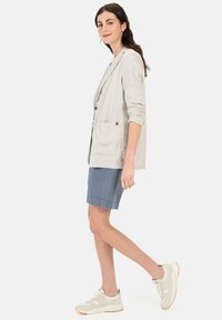 Lichtgrijze linnen blazer met opgerolde mouwen, enkele knoopsluiting en frontzakken, gecombineerd met een blauwe rok en beige sneakers.