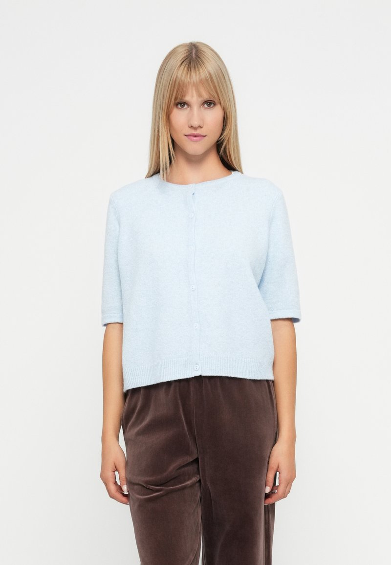 Ανοιχτό μπλε κοντομάνικο κάρντιγκ φτιαγμένο από μαλακό υλικό, με στρογγυλή λαιμόκοψη και κλείσιμο με κουμπιά μπροστά.