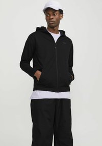 Schwarze Zip-Hoodie aus weichem Stoff mit langen Ärmeln, Seitentaschen und einer flachen Kapuze, über einem weißen T-Shirt getragen, kombiniert mit schwarzen Hosen.