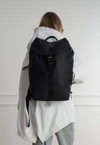 Zaino nero realizzato in materiale resistente, con chiusura roll-top, fibbia frontale e tasche laterali. Accentuato da un logo.