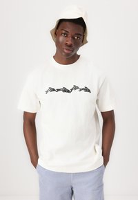 Weißes Baumwoll-T-Shirt mit einem schwarzen grafischen Druck von stilisierten Delfinen. Das Model trägt hellblaue gestreifte Shorts und einen beigen Bucket Hat.