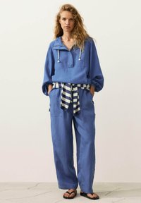 Femme aux longs cheveux bouclés portant un sweat à capuche bleu ample, un pantalon large assorti, un pull rayé noué à la taille et des sandales noires.