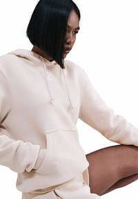 Felpa Nike beige chiara con cappuccio, caratterizzata da un cordino, tasca a kangaroo e polsini a costine. Tessuto morbido con design minimalista e dettaglio del logo.