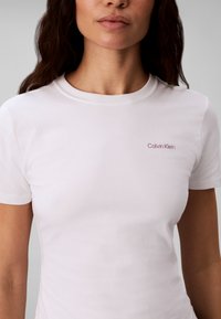 Γυναίκα που φοράει ένα απλό λευκό T-shirt Calvin Klein με κοντά μανίκια και στρογγυλή λαιμόκοψη, ορατή από το λαιμό έως τη μέση.