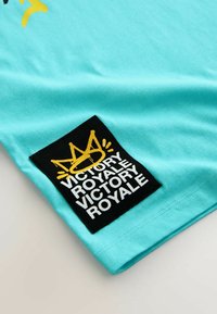 Tricou din bumbac turcoaz cu un eticheta neagră pe care sunt scrise cu alb cuvintele "VICTORY ROYALE" și un grafic cu o coroană galbenă. Textură moale.