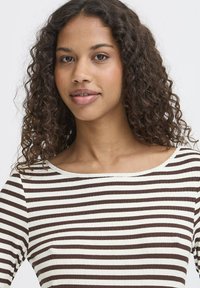 Brun og hvid stribet top med ribbet tekstur og bred halsudskæring. Krøllet hår rammer ansigtet, med et naturligt makeup-look.