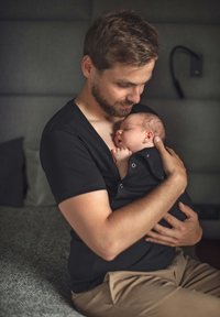 Sort stof bæresele til baby, båret af en mand, som holder en svøbt baby. Blød tekstur, V-hals design, neutral baggrund i lys grå nuancer.