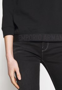Černá mikina s širokým lemem v pase s nápisem „EMPORIO ARMANI“ v decentním tónu. Dlouhé rukávy a hladká, měkká textura. Nošena s černými džíny.