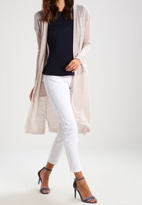 Ljusbeige lång cardigan med öppen framtill, kombinerad med en mörkblå peplumtopp och vita slim fit-byxor, med klackar som har ankelsnörning.