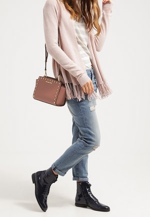Kobieta w jasnoróżowym kardiganie z frędzlami, w paskowanej bluzce, podartych, podwiniętych dżinsach, czarnych błyszczących butach na sznurowadła oraz z brązową torebką na ramię z ćwiekami.