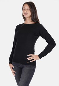 Maglione nero a maniche lunghe in tessuto a maglia, con scollo rotondo e un piccolo logo vicino all'orlo. Indossato con jeans scuri.