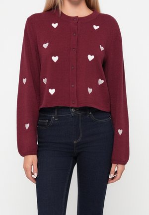 Mujer vestida con un cárdigan corto color granate con patrones de corazones blancos y jeans de cintura alta azul oscuro, posando frente a un fondo liso.
