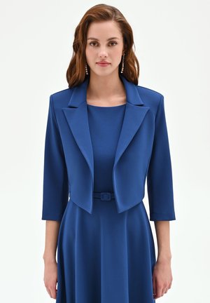 Donna con capelli castani ondulati indossa un vestito blu con cintura e un blazer corto abbinato con maniche a tre quarti e orecchini pendenti.