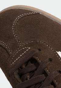 Chaussure en daim marron avec des coutures et des lacets blancs. Comprend des œillets renforcés et des détails de surface texturée pour une meilleure adhérence.
