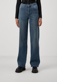 High-waisted, loose-fit blue denim jeans with five-pocket design, faded texture, and belt loops, paired with black chunky boots.