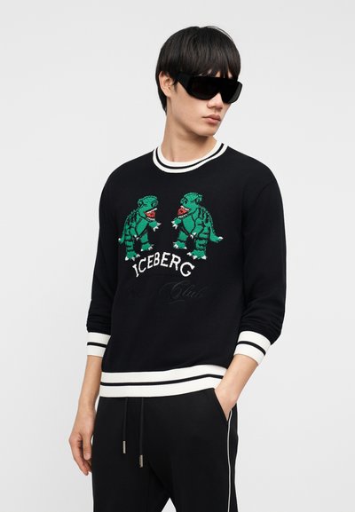 Νέος άνδρας που φοράει υπερμεγέθη μαύρα γυαλιά ηλίου και μαύρο πουλόβερ με πράσινους δεινόσαυρους και μανσέτες και τελείωμα με λευκές ρίγες.