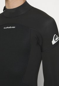 Muta in neoprene nera con un design aderente. Presenta cuciture visibili e un logo Quiksilver sulla spalla destra.