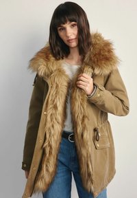 Kurtka parka z kapturem w oliwkowym kolorze, z futrzanym kołnierzem i podszewką. Wykonana z solidnego materiału, z bocznymi kieszeniami i regulowanym paskiem w talii.