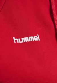 Tissu rouge avec logo blanc de la marque hummel imprimé sur la surface, montrant une partie d’un cordon tressé sur le côté.