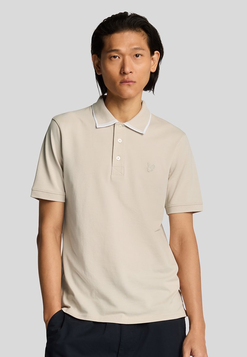 Polo shirt beige con maniche corte, rifinitura del colletto bianca e patta con tre bottoni. Presenta logo ricamato sul petto. Tessuto morbido.
