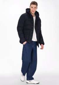 Tengerészkék bélelt dzseki kapucnival, cipzáras elülső résszel és gombos záródással; két elülső zsebével. Laza kék farmerrel és fehér tornacipővel viselve.