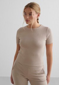 Ribbet beige t-shirt med korte ærmer og rund hals, der har et slankt, tætsiddende design og en subtil logo-detalje ved kanten.