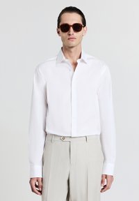 Chemise blanche à manches longues avec col boutonné, associée à un pantalon beige. Des lunettes de soleil brun foncé complètent la tenue. Texture unie, coupe ajustée.