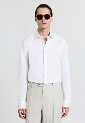 Camicia bianca a maniche lunghe con colletto button-down, abbinata a pantaloni beige. Gli occhiali da sole marroni scuri completano l'outfit. Tessuto semplice, vestibilità su misura.