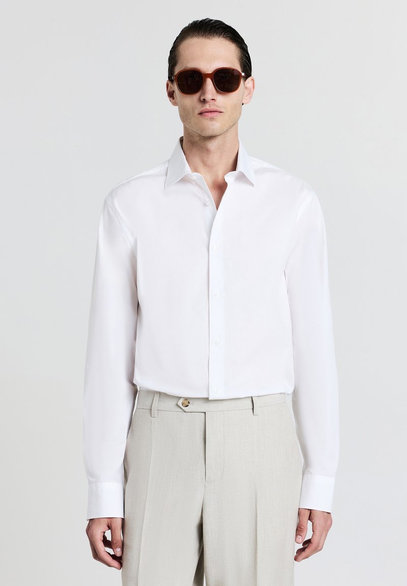 Chemise blanche à manches longues avec col boutonné, associée à un pantalon beige. Des lunettes de soleil brun foncé complètent la tenue. Texture unie, coupe ajustée.