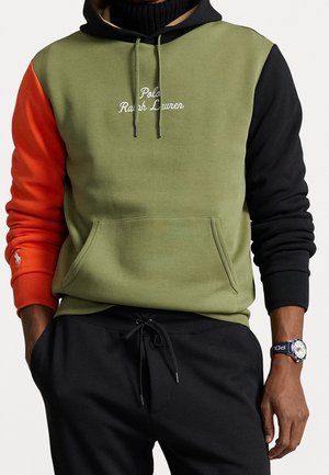 Mężczyzna w zielonej bluzie z kapturem z pomarańczowym lewym rękawem, czarnym prawym rękawem i napisem "Polo Ralph Lauren", w czarnych spodniach i z zegarkiem na nadgarstku.