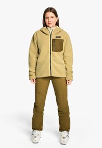 Beige Fleecejacke mit Reißverschluss, großer Kapuze und olivgrünem Brusttasche. Olivgrüne Hose mit tailliertem Schnitt und Logodetail.