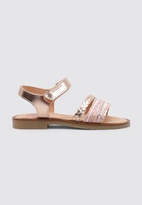 Sandalias metálicas en tono rosa oro con una correa trenzada, que cuentan con una banda tejida de varios colores y una correa ajustable en el tobillo. Diseño de suela plana.
