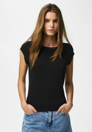 Ung kvinne med langt brunt hår som har på seg en enkel svart ermeløs topp og blå jeans, hender i lommer, nøytral uttrykk.