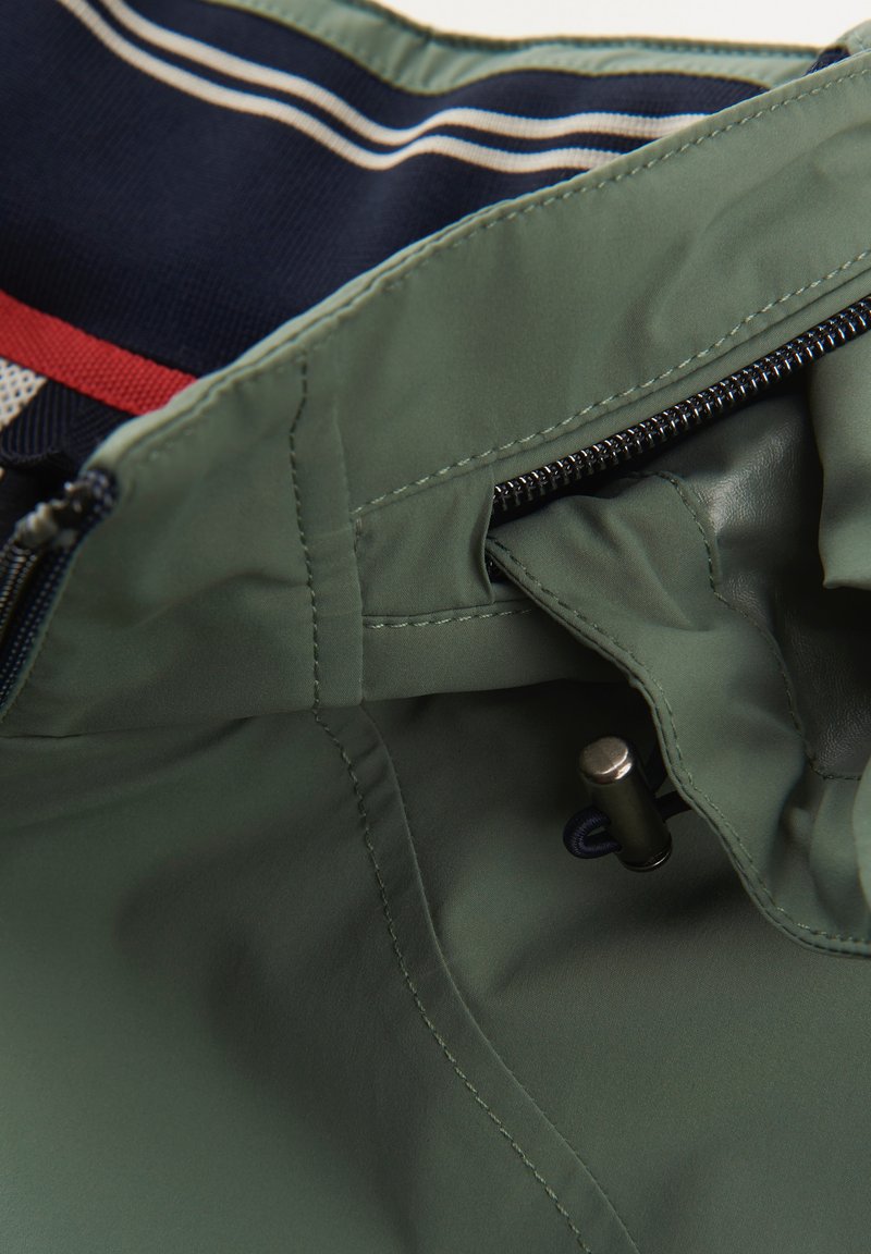 Intérieur d'une veste imperméable verte avec une doublure rayée bleue et blanche, comportant une fermeture à glissière et un cordon de serrage pour l'ajustement.