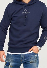 Navyn blå hoodie gjord av bomullsblandning, med en dragsko i huva, ribbade ärmslut och en logotyp på bröstet och ärmen. Buren över slitna jeans.