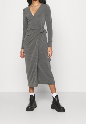 Femme portant une robe midi portefeuille grise à manches longues avec un nœud sur le côté, des chaussettes blanches et des bottines noires épaisses, debout devant un fond uni.