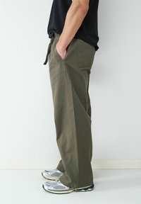 Olijfgroene broek met een ontspannen pasvorm, een koordsluiting in de taille, zijzakken en een gladde textuur, gecombineerd met zilveren sportschoenen.