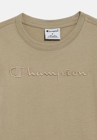Ανοιχτό καφέ βαμβακερό μπλουζάκι με στρογγυλή λαιμόκοψη και κεντημένο λογότυπο "Champion" σε πιο σκούρο καφέ, με απαλό υφή.