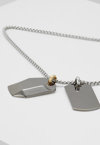 Metal halskæde med to vedhæng: det ene har en hexagonal form med en sølvfinish og indgraveret tekst; det andet er et rektangulært sølvmærke.