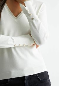 Bílý hladký top s dlouhým rukávem a výstřihem do V, s vlnitým ozdobným lemem a třemi zlatými knoflíky na manžetě. Přiléhavý střih.