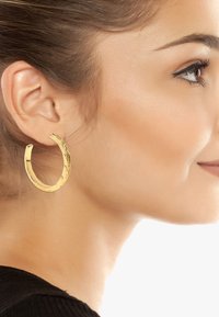 Goldene Creolen mit einem gedrehten Design, mittlere Größe, glatte Oberfläche, reflektieren das Licht, getragen am Ohr eines Models.