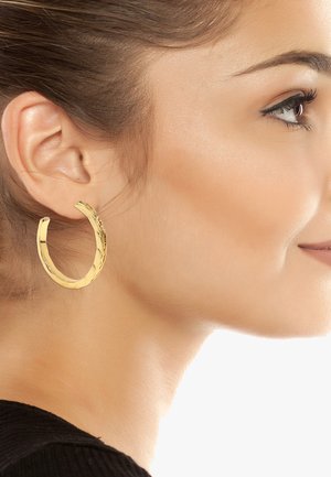 Pendientes de aro de oro con un diseño retorcido, de tamaño mediano, acabado suave, que refleja la luz, usados en la oreja de un modelo.