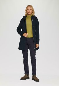 Manteau en mélange de laine bleu marine avec une capuche, fermé par des boutons argentés ; porté sur un pull à col roulé vert et un pantalon sombre ; bottines brunes à la cheville.