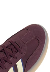 Tamno crvena atletska cipela sa antilop gornjim dijelom, sa krem i plavim bočnim prugama, okruglim vezicama i teksturiranom gumom na potplatu.