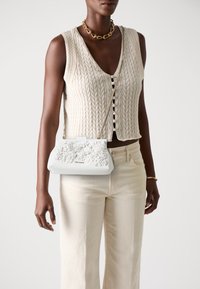 Borsa a mano azzurro chiaro con motivo floreale in rilievo, tracolla dorata, forma rettangolare. Indossata con un gilet di maglia crema e pantaloni a gamba larga beige.