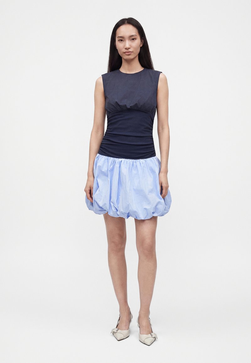 Femme aux longs cheveux foncés portant un haut sans manches bleu marine et une jupe bulle bleu clair, debout, les bras détendus le long du corps, sur un fond uni.