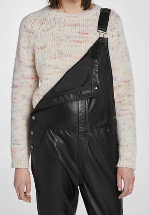 Person wearing a white speckled knit sweater and black leather overalls with one strap undone and folded down.