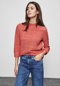 Maglione rosso effetto texture con maniche lunghe e collo alto, abbinato a jeans in denim blu. L'outfit presenta una vestibilità comoda e uno stile informale.