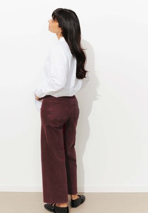 Femme aux longs cheveux noirs portant une chemise blanche, un pantalon large violet et des chaussures noires à clous, debout de profil contre un mur blanc.