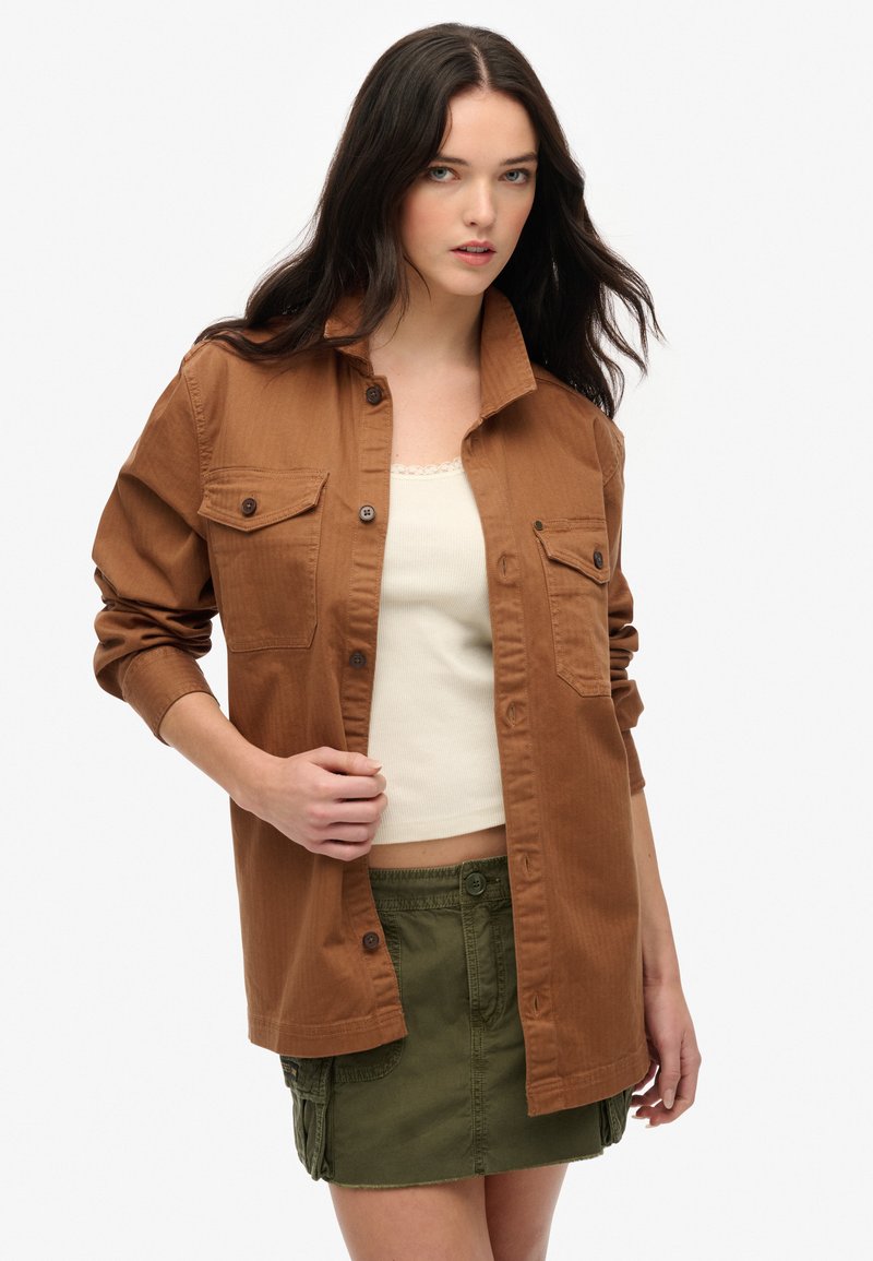 Brązowa koszula z guzikami, z dwiema kieszeniami na piersi, podwiniętymi rękawami i prostym kołnierzykiem, noszona na kremowej koszulce na ramiączkach i zielonej spódnicy.